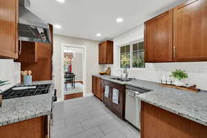 Kitchen featuring decorative backsplash, light stone counters, wall chimney range hood, brown cabinetry, and recessed lighting