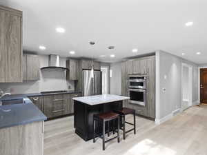 Kitchen with a breakfast bar area, light wood-type flooring, appliances with stainless steel finishes, hanging light fixtures, and wall chimney exhaust hood