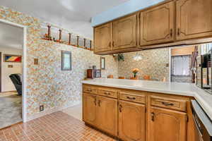 Kitchen looking into dining. Light countertops, wallpapered walls, brick patterned flooring, and brown cabinetry