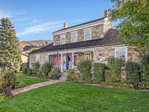 View of front facade featuring a front lawn, stone siding, covered porch, a high end roof, and a chimney