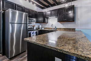 Kitchen featuring appliances with stainless steel finishes, dark stone countertops, a peninsula, dark wood-style flooring, and dark cabinetry