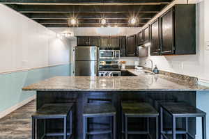 Kitchen featuring appliances with stainless steel finishes, light stone counters, dark wood-type flooring, a peninsula, and a kitchen breakfast bar
