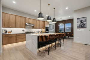Kitchen with hanging light fixtures, an island with sink, light stone counters, a kitchen breakfast bar, and under cabinet range hood