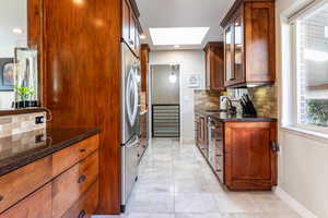 Kitchen featuring a skylight, backsplash, dark stone counters, glass insert cabinets, and brown cabinets