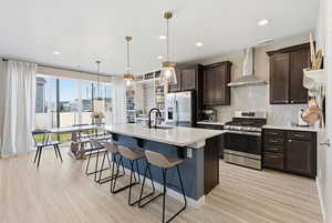 Kitchen with appliances with stainless steel finishes, wall chimney exhaust hood, dark brown cabinetry, a kitchen breakfast bar, and an island with sink