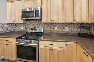 Kitchen with appliances with stainless steel finishes, light brown cabinetry, decorative backsplash, and dark countertops