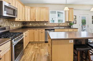 Kitchen with light brown cabinetry, appliances with stainless steel finishes, decorative backsplash, dark countertops, and a breakfast bar area