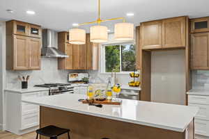 Kitchen featuring glass insert cabinets, hanging light fixtures, decorative backsplash, brown cabinetry, and wall chimney range hood