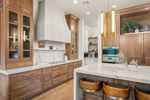 Kitchen with open shelves, brown cabinetry, custom range hood, light stone countertops, and recessed lighting