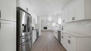 Kitchen with a glass covered fireplace, stainless steel appliances, white cabinetry, light wood-type flooring, and light stone counters