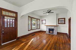 Unfurnished living room featuring arched walkways, dark wood-style flooring, a brick fireplace, and a ceiling fan