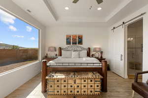 Bedroom with a barn door, light wood finished floors, a raised ceiling, ceiling fan, and connected bathroom