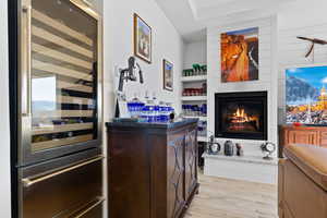 Indoor wet bar with wine cooler, light wood-style flooring, dark brown cabinets, a glass covered fireplace, and dark countertops