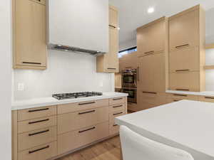 Kitchen with light brown cabinets, custom exhaust hood, light wood-type flooring, and appliances with stainless steel finishes