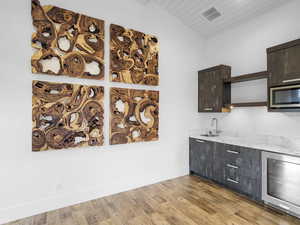 Bar area featuring beverage cooler, open shelves, lofted ceiling, stainless steel microwave, and light wood-type flooring