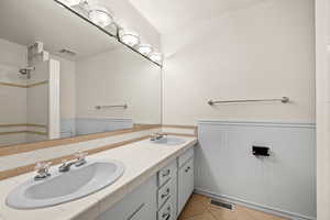 Full bathroom featuring a wainscoted wall, double vanity, tiled shower, and light tile patterned floors