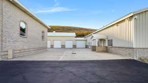 View of property exterior featuring brick siding, driveway, a view of trees, a metal roof, and an outdoor structure