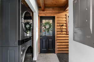 Laundry room with rustic walls, electric panel, a wooden ceiling with exposed beams, and stacked washer / dryer