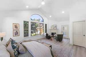 Bedroom featuring light wood-style floors, a glass covered fireplace, ceiling fan, high vaulted ceiling, and access to outside