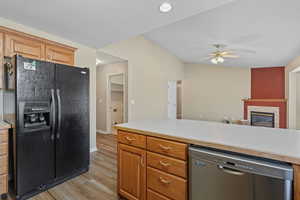 Kitchen featuring black refrigerator with ice dispenser, light countertops, stainless steel dishwasher, a fireplace, and light wood-style flooring