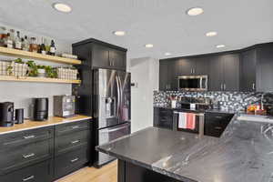Kitchen with stainless steel appliances, open shelves, dark cabinets, light wood-style floors, and tasteful backsplash