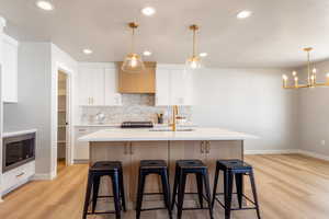 Kitchen featuring white cabinetry, backsplash, light wood-style floors, decorative light fixtures, and a kitchen island with sink