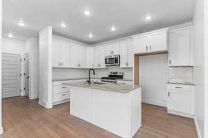 Kitchen with white cabinets, appliances with stainless steel finishes, backsplash, light wood-type flooring, and a center island with sink