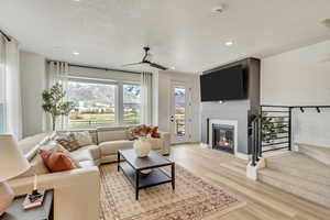 Living room with a ceiling fan, light wood-type flooring, a glass covered fireplace, stairway, and recessed lighting