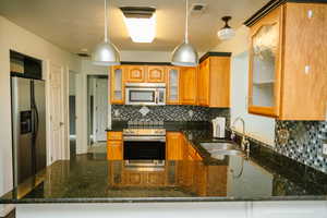 Kitchen with glass insert cabinets, tasteful backsplash, dark granite countertops, hanging light fixtures, and stainless steel appliances