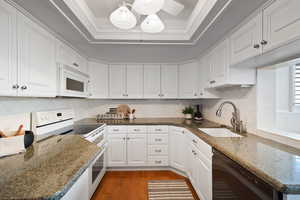 Kitchen featuring a raised ceiling, dark stone countertops, backsplash, white appliances, and ornamental molding