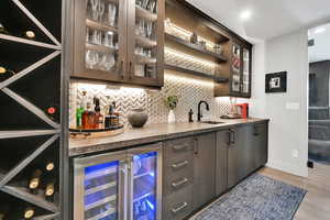 Indoor bar with dark brown cabinetry, backsplash, glass insert cabinets, beverage cooler, and light wood-style flooring
