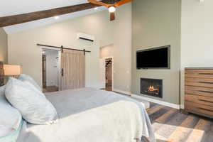 Bedroom with dark wood-style flooring, a glass covered fireplace, a barn door, high vaulted ceiling, and ceiling fan