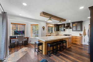 Kitchen featuring glass insert cabinets, a textured ceiling, tasteful backsplash, dark wood finished floors, and wall chimney range hood
