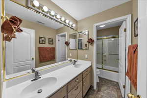 Bathroom featuring double vanity, a textured ceiling, shower / bath combination with glass door, and dark stone finish flooring