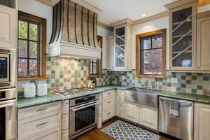 Kitchen featuring appliances with stainless steel finishes, custom exhaust hood, decorative backsplash, dark wood-style floors, and glass insert cabinets