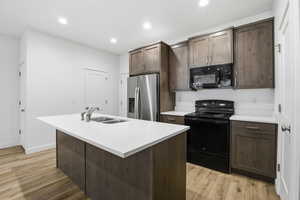 Kitchen with dark brown cabinets, black appliances, light wood-type flooring, an island with sink, and recessed lighting