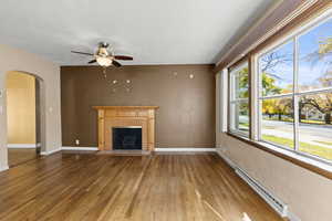 Unfurnished living room with a baseboard heating unit, arched walkways, wood-type flooring, a brick fireplace, and ceiling fan