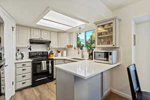 Kitchen with black range with electric cooktop, decorative light fixtures, stainless steel microwave, light wood-style flooring, and glass insert cabinets