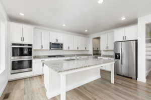 Kitchen featuring stainless steel appliances, white cabinetry, light stone countertops, a kitchen island with sink, and light wood-style floors