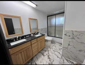 Bathroom with double vanity, light marble finish floors, tile walls, shower / bath combination with glass door, and a wainscoted wall