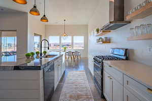 Kitchen featuring stainless steel gas stove, wall chimney range hood, open shelves, hanging light fixtures, and white cabinets