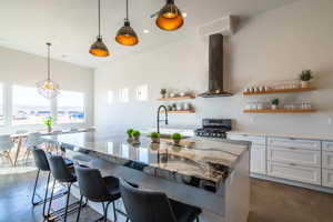 Kitchen featuring open shelves, pendant lighting, light stone countertops, white cabinetry, and wall chimney range hood