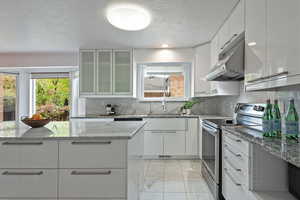 Kitchen featuring stainless steel range with electric cooktop, white cabinets, glass insert cabinets, light stone countertops, and a textured ceiling