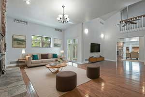 Living room with wood-type flooring, a stone fireplace, ceiling fan, high vaulted ceiling, and a chandelier