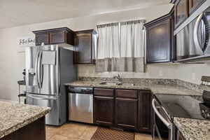 Kitchen featuring stainless steel appliances, dark brown cabinetry, light tile patterned flooring, light stone counters, and a textured ceiling