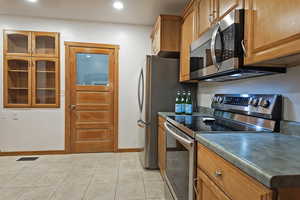 Kitchen with appliances with stainless steel finishes, glass insert cabinets, brown cabinetry, light tile patterned floors, and dark countertops