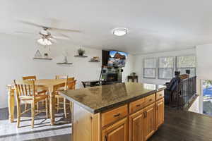 Kitchen with a warm lit fireplace, a center island, dark wood-style flooring, dark stone countertops, and ceiling fan