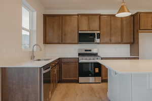 Kitchen with stainless steel appliances, light wood-style floors, pendant lighting, light stone counters, and brown cabinetry