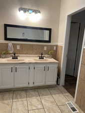 Full bath with a textured wall, double vanity, tile walls, wainscoting, and light tile patterned flooring