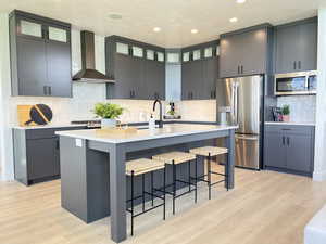 Kitchen with a kitchen breakfast bar, stainless steel appliances, a kitchen island with sink, wall chimney exhaust hood, and backsplash
